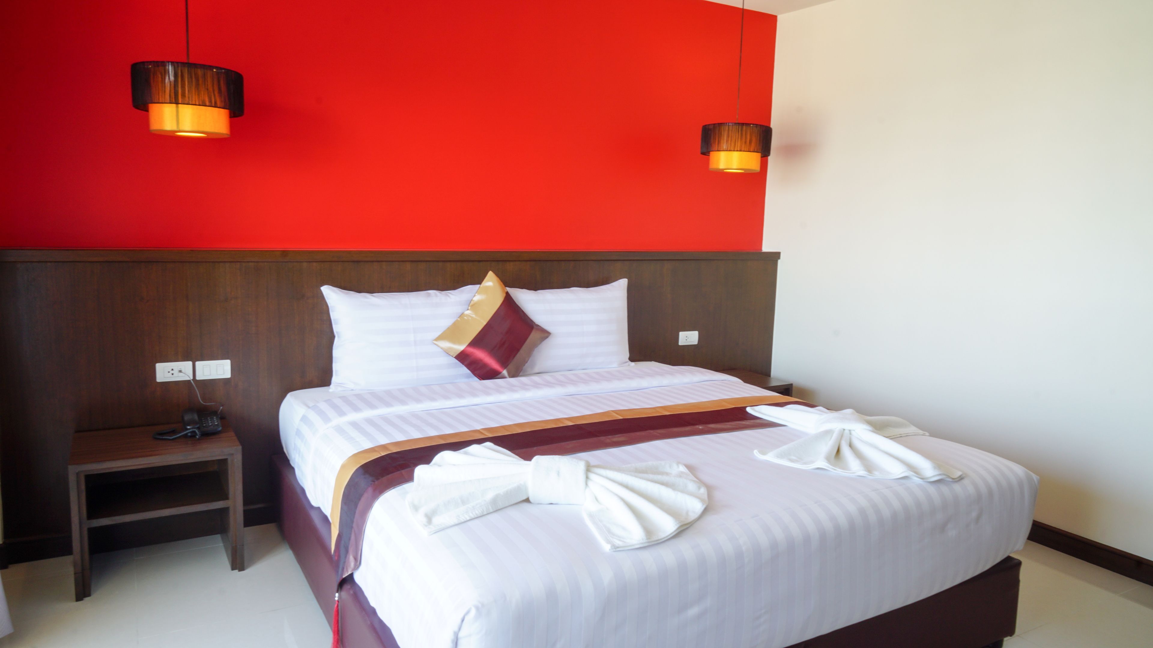 Une chambre d’hôtel avec un grand lit, deux tables de chevet, un mur d’accent rouge et deux lampes suspendues.