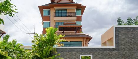 A modern multi-story building with a unique tiered roof design, surrounded by lush greenery and a paved walkway.