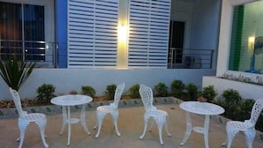 Uma área de pátio com cadeiras e mesas de metal branco, um pequeno jardim com plantas e um edifício com persianas brancas e um lustre.