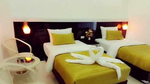 Quarto de hotel com duas camas, cada uma com colcha amarela e almofadas brancas. Há uma cadeira branca e uma pequena mesa com um vaso de flores.