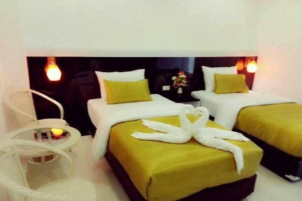 Una habitación de hotel con dos camas, cada una con cubrecama amarillo y almohadas blancas. Hay una silla blanca y una mesita con un jarrón de flores.