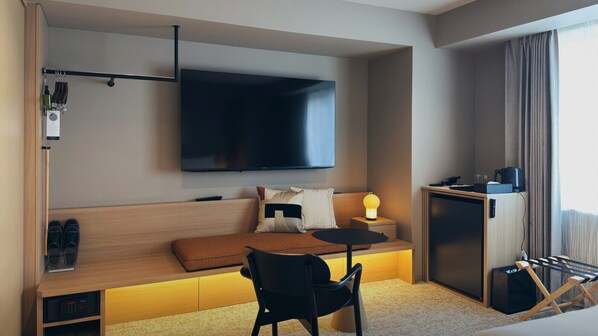 Una habitación de hotel moderna con un televisor de pantalla plana grande montado en la pared, una zona de estar acogedora con un sofá y almohadas, una pequeña mesa redonda con una lámpara, un escritorio con una computadora y una cafetera, y un zapatero.