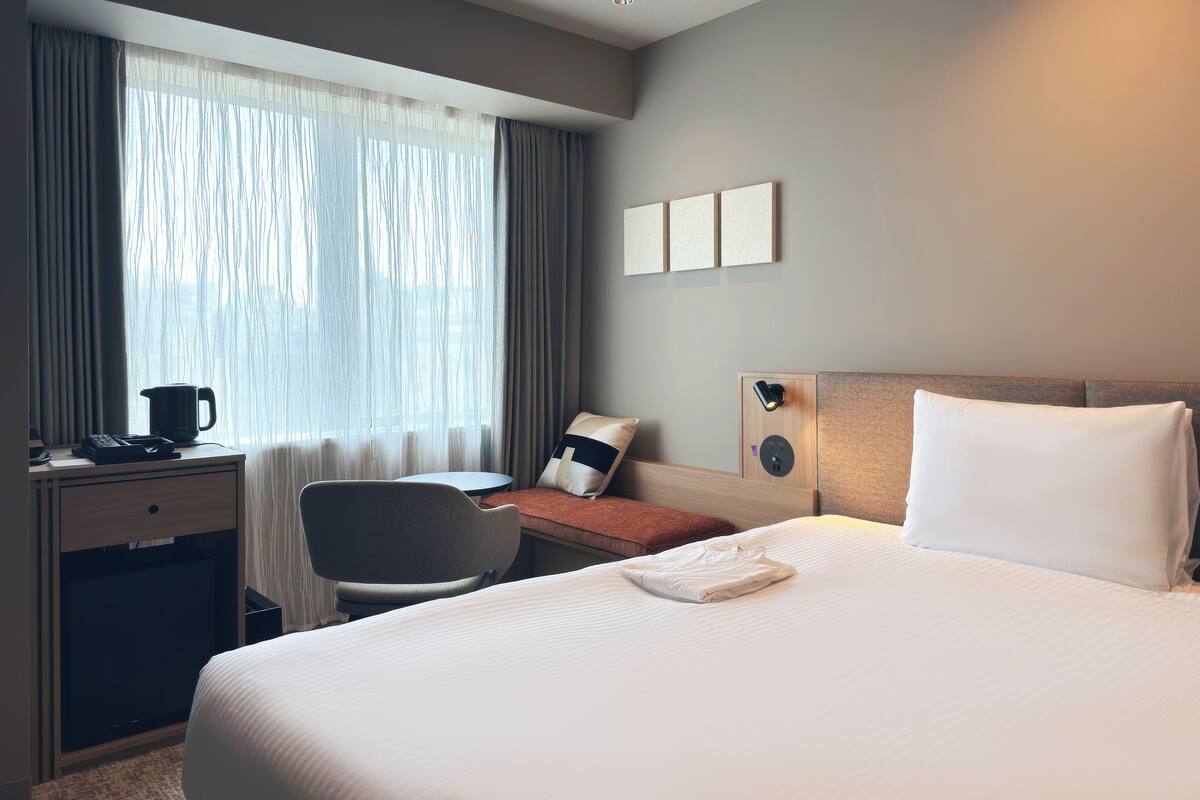 A hotel room with a bed, a desk with a coffee maker, a chair, and a window with curtains.