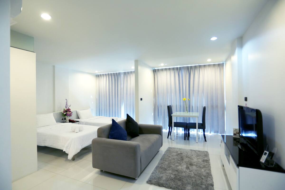 Un salon moderne et bien éclairé, comprenant un canapé, une table à manger avec des chaises, un téléviseur et un lit avec une couette blanche.