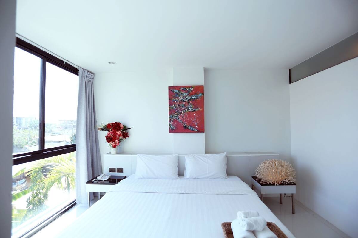 Une chambre d’hôtel moderne avec un grand lit, une table de chevet avec un téléphone, un vase de fleurs, un tableau au mur et une vue sur la ville par la fenêtre.