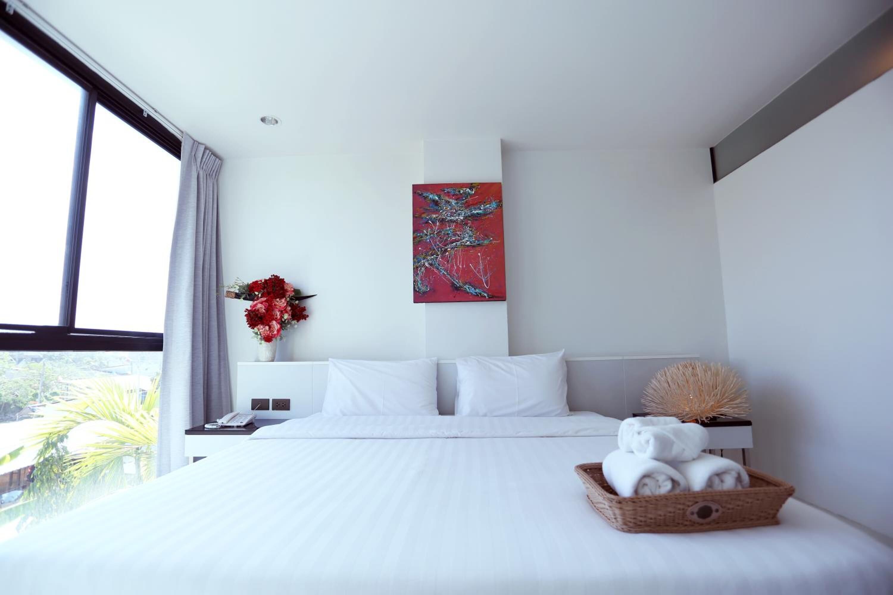 Une chambre d’hôtel moderne avec un grand lit, du linge de lit blanc, un tableau abstrait rouge, un vase rempli de fleurs rouges et un panier rempli de serviettes.