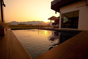 A swimming pool with a wooden deck, a covered seating area, and a view of mountains at sunset. - The Vista Pool Villa (Tha Muang)
