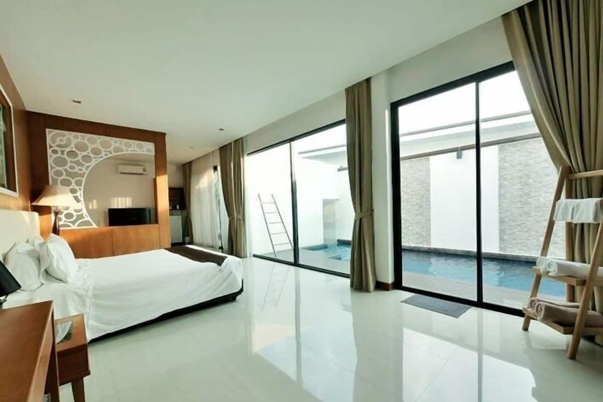 A modern hotel room with a large bed, a desk, a chair, a TV, and a view of an infinity pool through large windows.