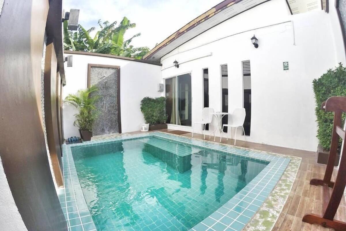 A rectangular swimming pool with tiled edges, surrounded by a white wall with a glass door and windows. There are white chairs and a small table near the pool, and a wooden chair is visible on the right side.