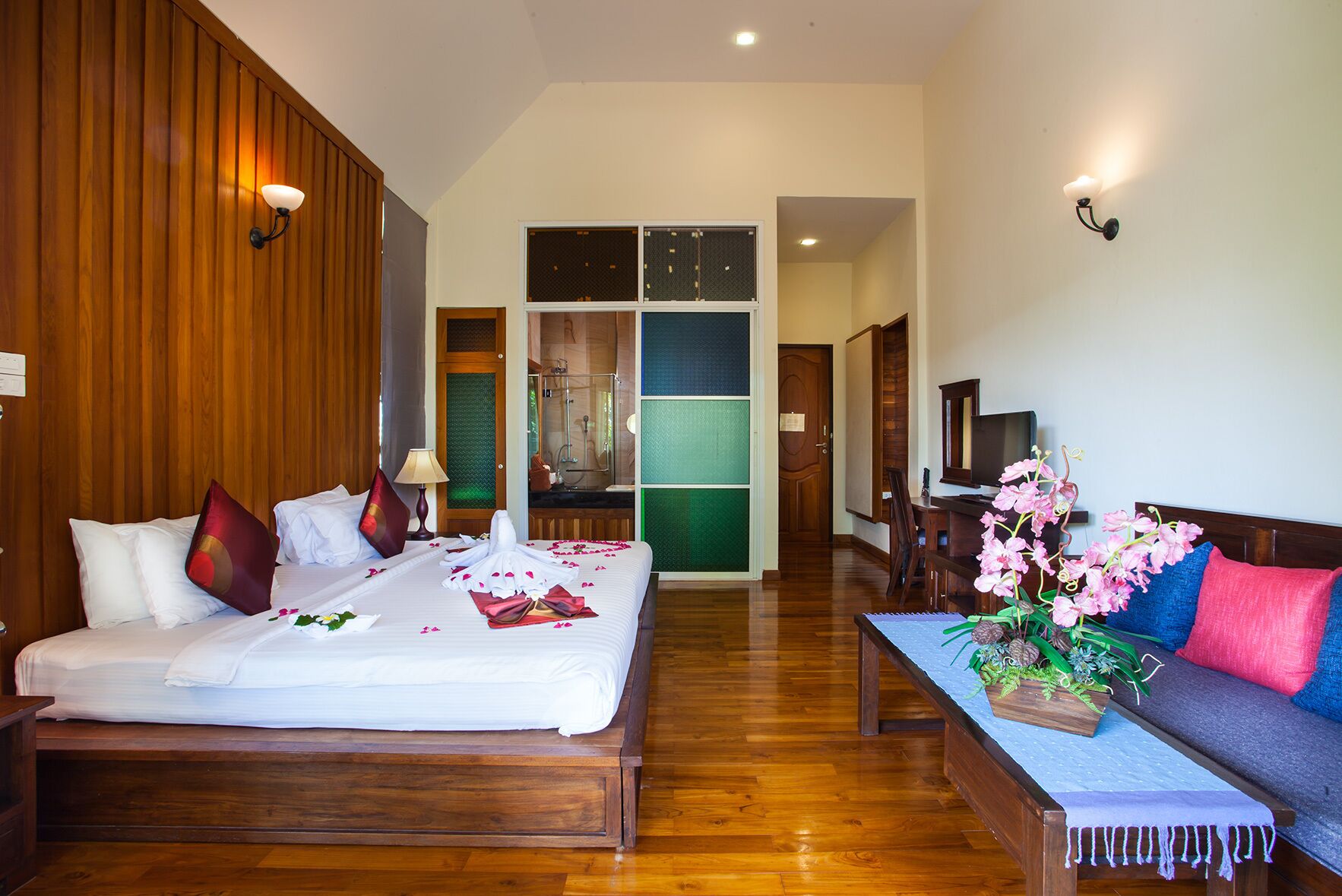 Habitación de hotel con cama de madera, sofá, escritorio y baño visible a través de una puerta de cristal.