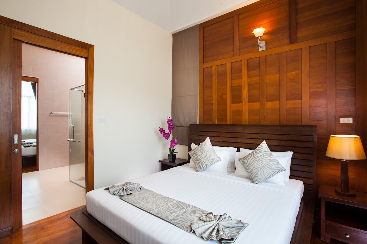 A hotel room with a wooden headboard, a bed with white linens, a nightstand with a lamp, and a bathroom visible through an open door.