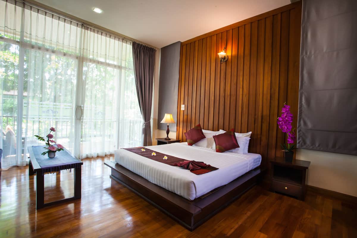 Um quarto espaçoso com uma cama grande, painéis de madeira nas paredes, uma mesa de cabeceira com luminária e vista para o verde pela janela.
