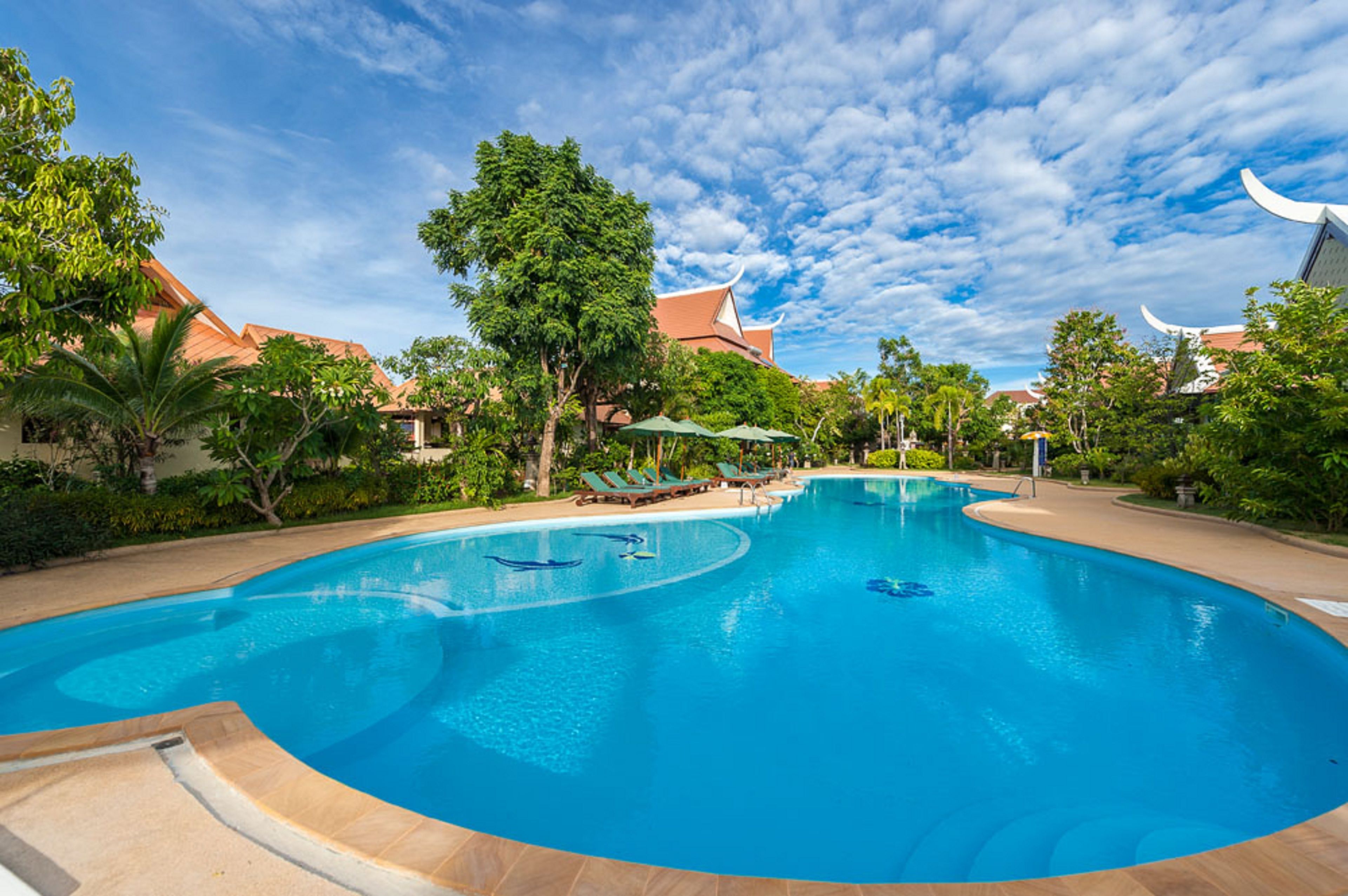 Uma piscina ampla e convidativa, com espreguiçadeiras e guarda-sóis, cercada por vegetação exuberante e arquitetura tradicional.