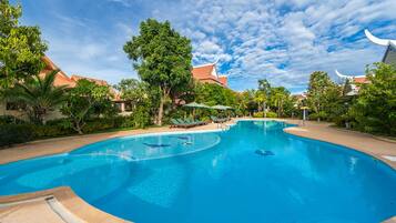 Uma piscina ampla e convidativa, com espreguiçadeiras e guarda-sóis, cercada por vegetação exuberante e arquitetura tradicional.