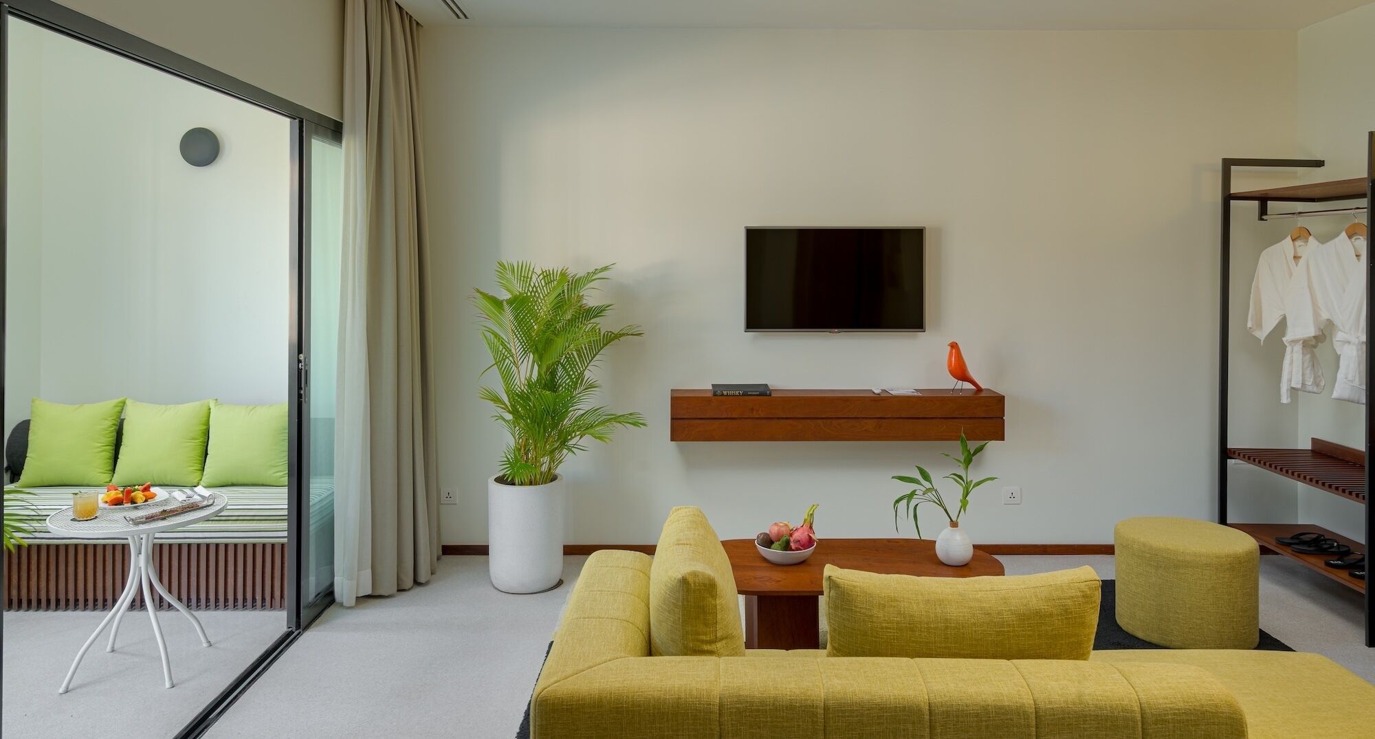A modern hotel room with a sofa, a dining table, a TV, a plant, and a clothing rack.
