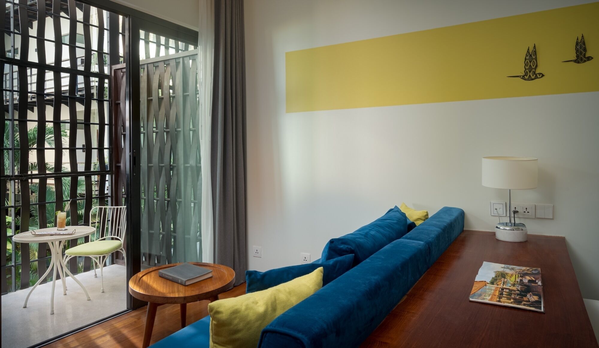 A room with a blue sofa, a small round table with a book, a lamp, and a view through a glass door.