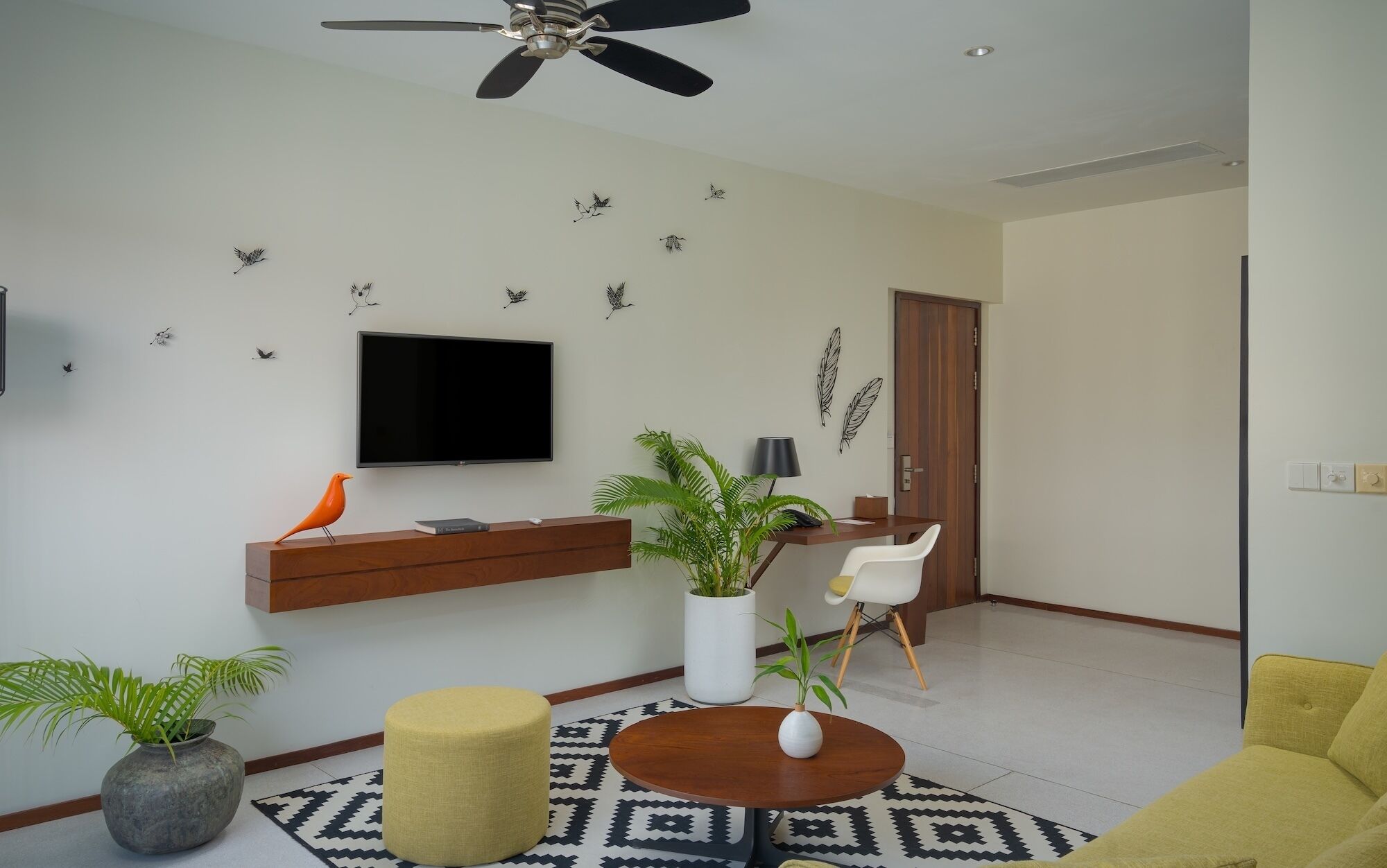 A living room with a flat-screen TV, a ceiling fan, a wooden TV stand, a round coffee table, a yellow ottoman, a white chair, and a patterned rug.