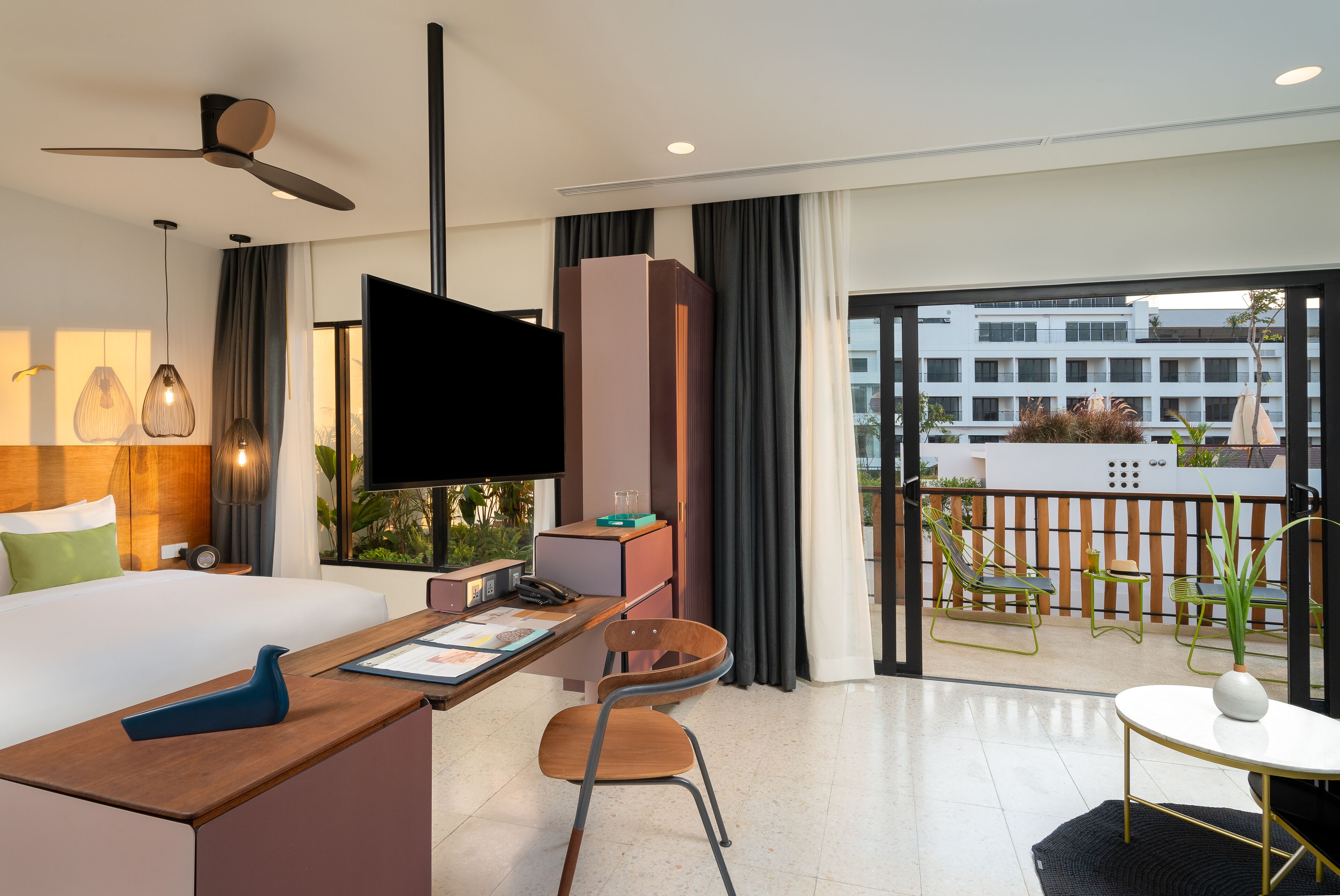 A modern hotel room with a bed, desk, chair, TV, and balcony with outdoor furniture.