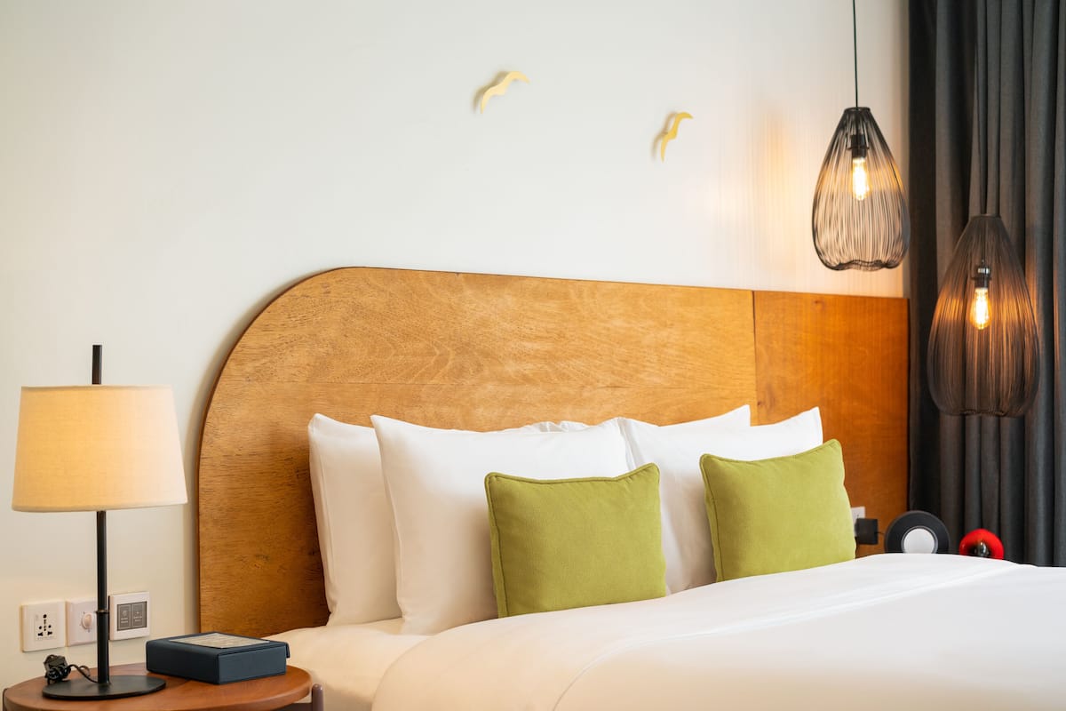 A hotel room with a wooden headboard, white bedding, green pillows, a bedside table with a lamp, and two pendant lights.