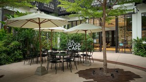 Een terras met tafels en stoelen, omringd door groen en parasols, met een bordje waarop 'The Flock Cafe' staat.