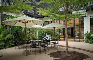 An outdoor seating area with tables and chairs, surrounded by greenery and umbrellas, with a sign reading 'The Flock Cafe'.
