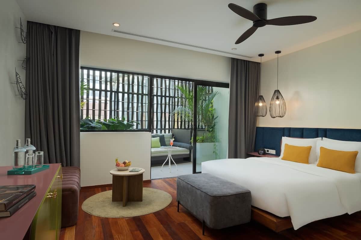A hotel room with a bed, a desk, a chair, a small table with a bowl of fruit, and a balcony with a view.