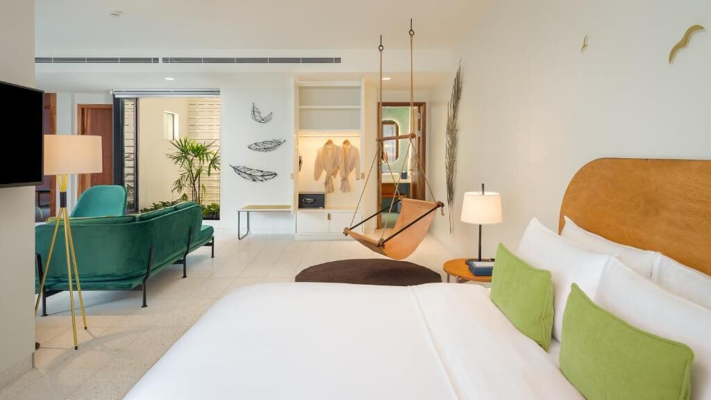 A modern hotel room with a large bed, a hanging chair, a green armchair, a TV, and a wardrobe.