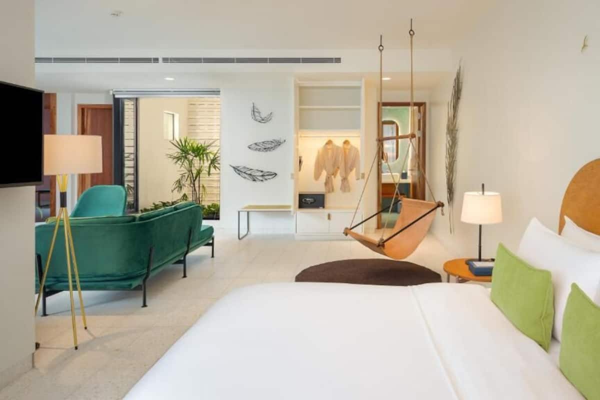 A modern hotel room with a large bed, a hanging chair, a green armchair, a TV, and a wardrobe.