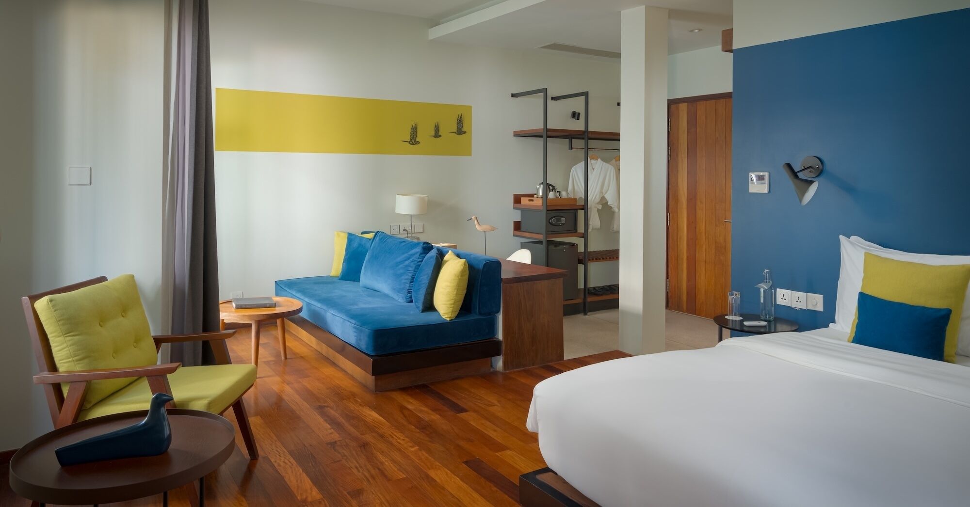 A hotel room with a bed, a blue sofa, a wooden chair, a small table, and a desk with a lamp.