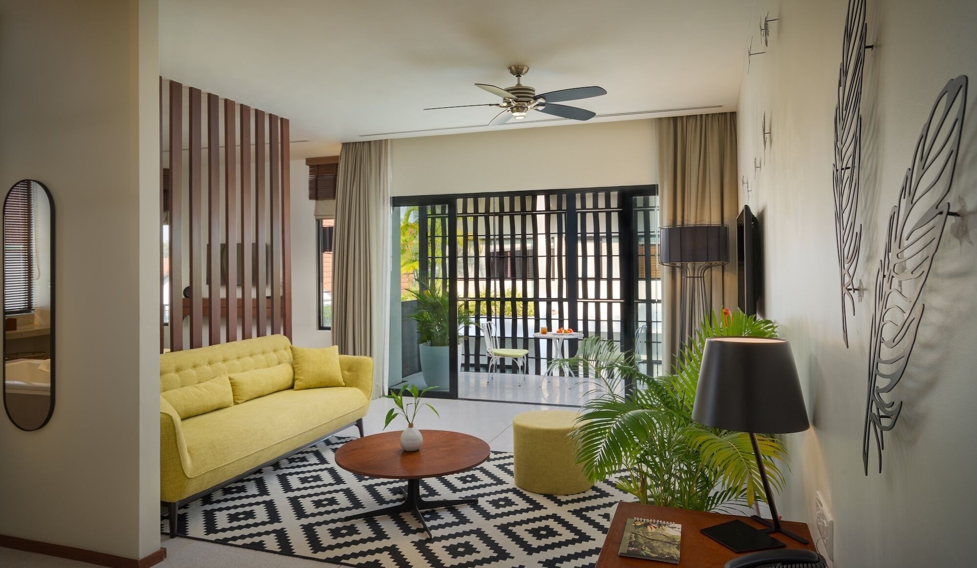 A living room with a yellow sofa, a round coffee table, a patterned rug, a potted plant, and a ceiling fan.