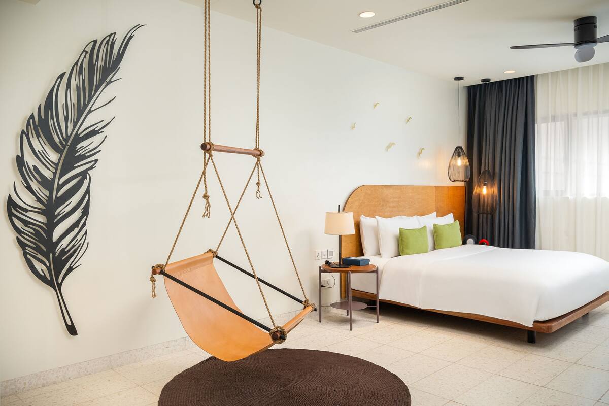 A modern hotel room with a large bed, a hanging chair, a nightstand, and a wall art piece.
