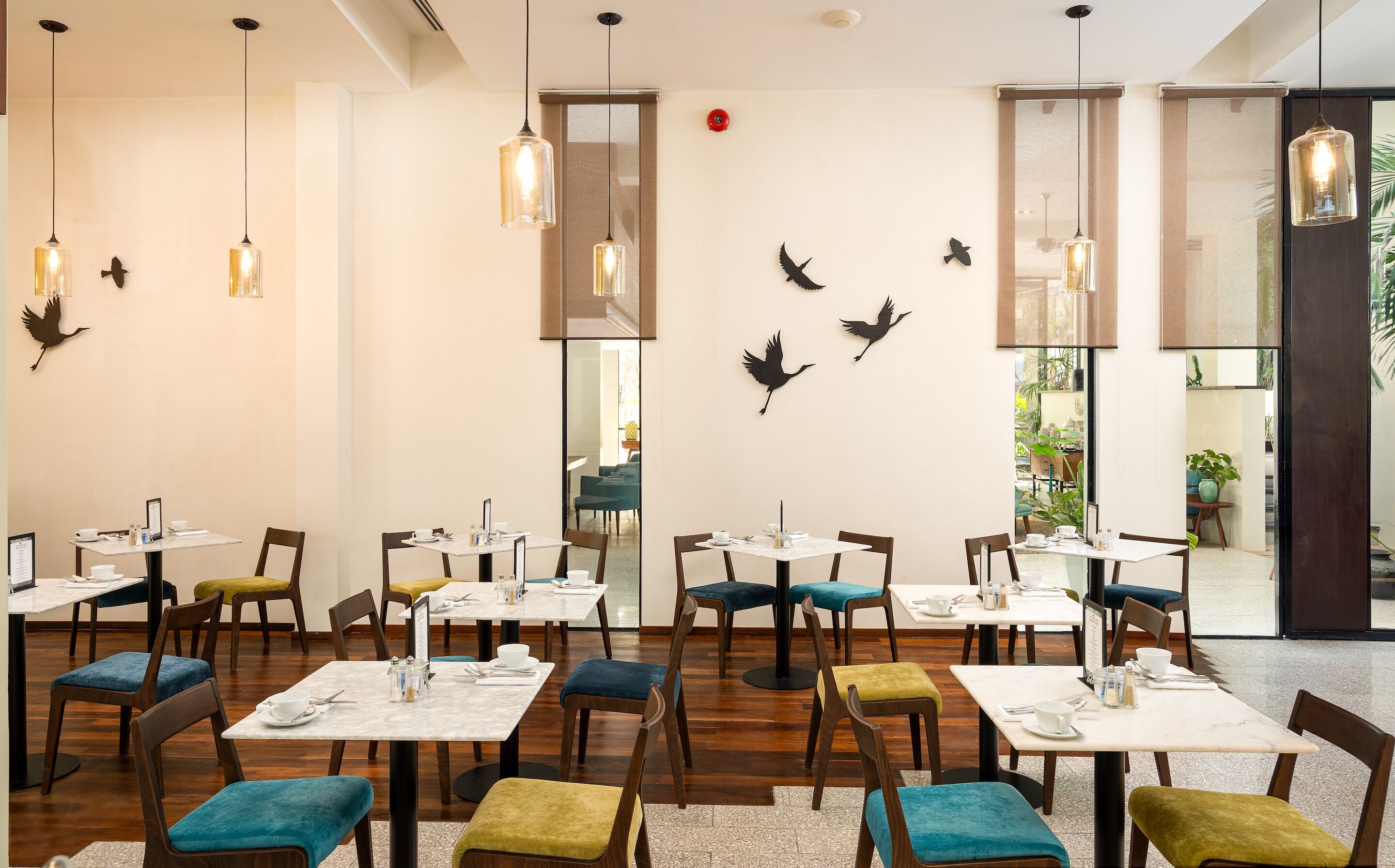 Un restaurant amb diverses taules i caires, algunes amb coixins de colors. Hi ha llums penjants i decoracions de parets amb motius d'ocells.