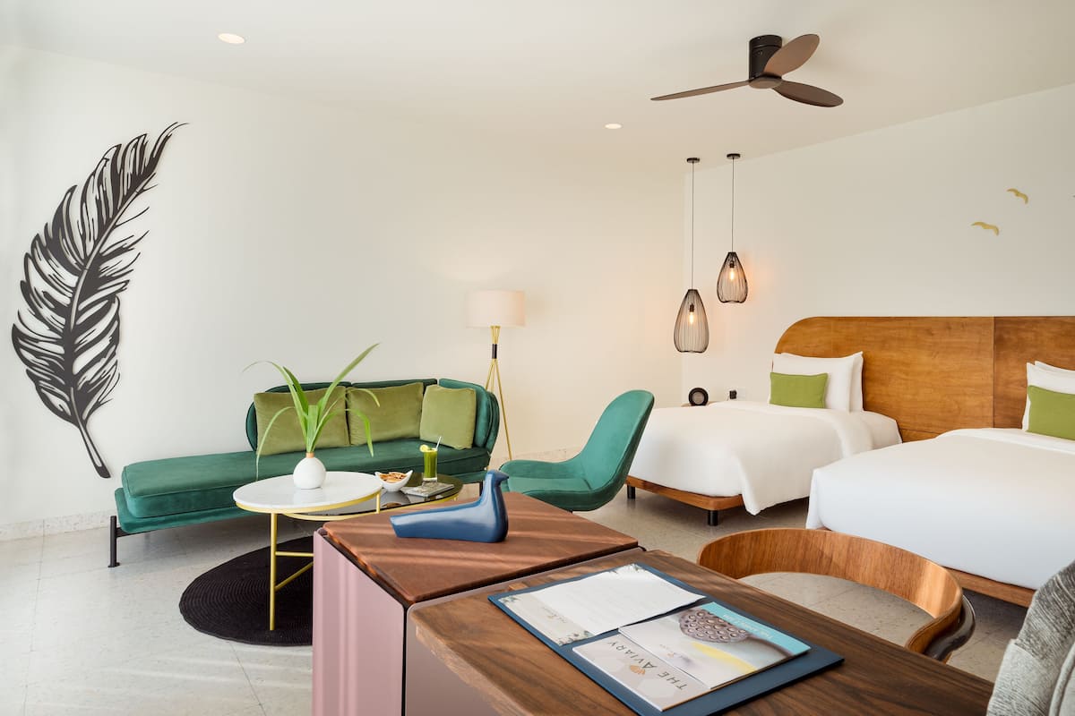 A hotel room with a bed, two chairs, a desk, a green sofa, a decorative leaf wall art, a ceiling fan, and a hanging light fixture.