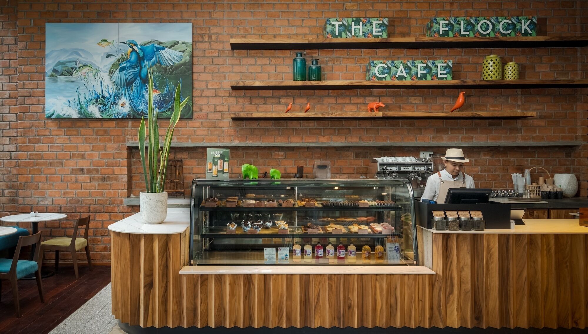 A cafe with a brick wall, a painting, shelves with items, a display case with pastries, and a barista.