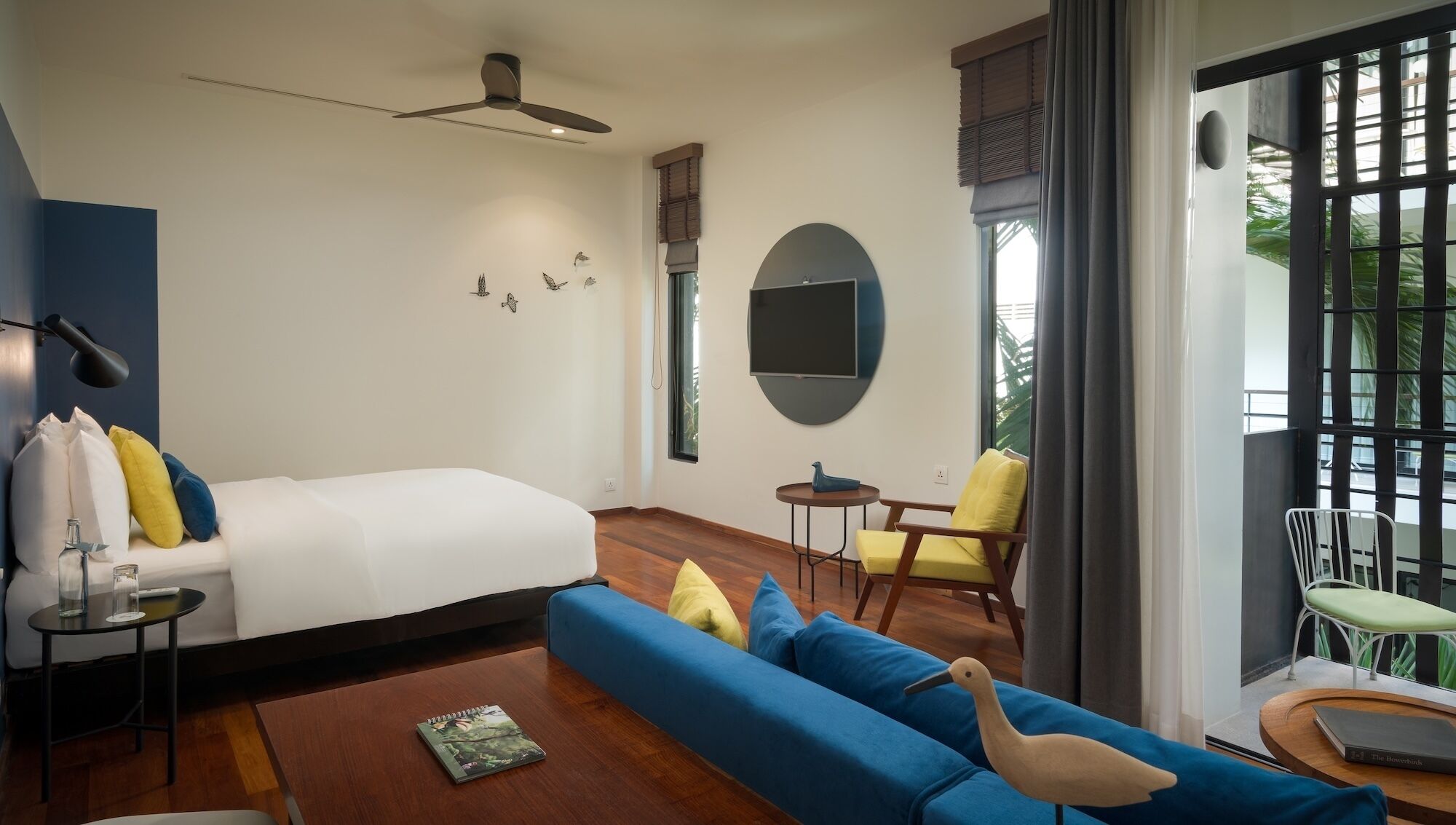 A modern hotel room with a bed, sofa, chairs, a TV, and a view of greenery.