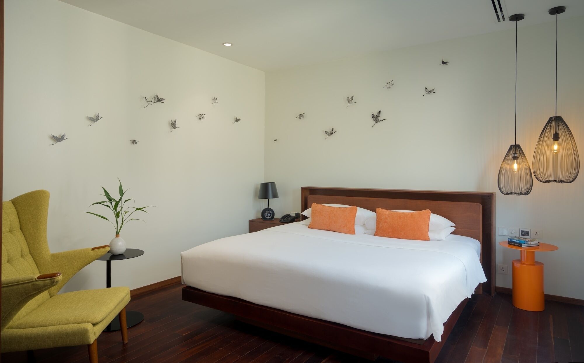 A hotel room with a bed, a chair, a small table, a lamp, wall art, and pendant lights.