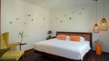 A hotel room with a bed, a chair, a small table, a lamp, wall art, and pendant lights.