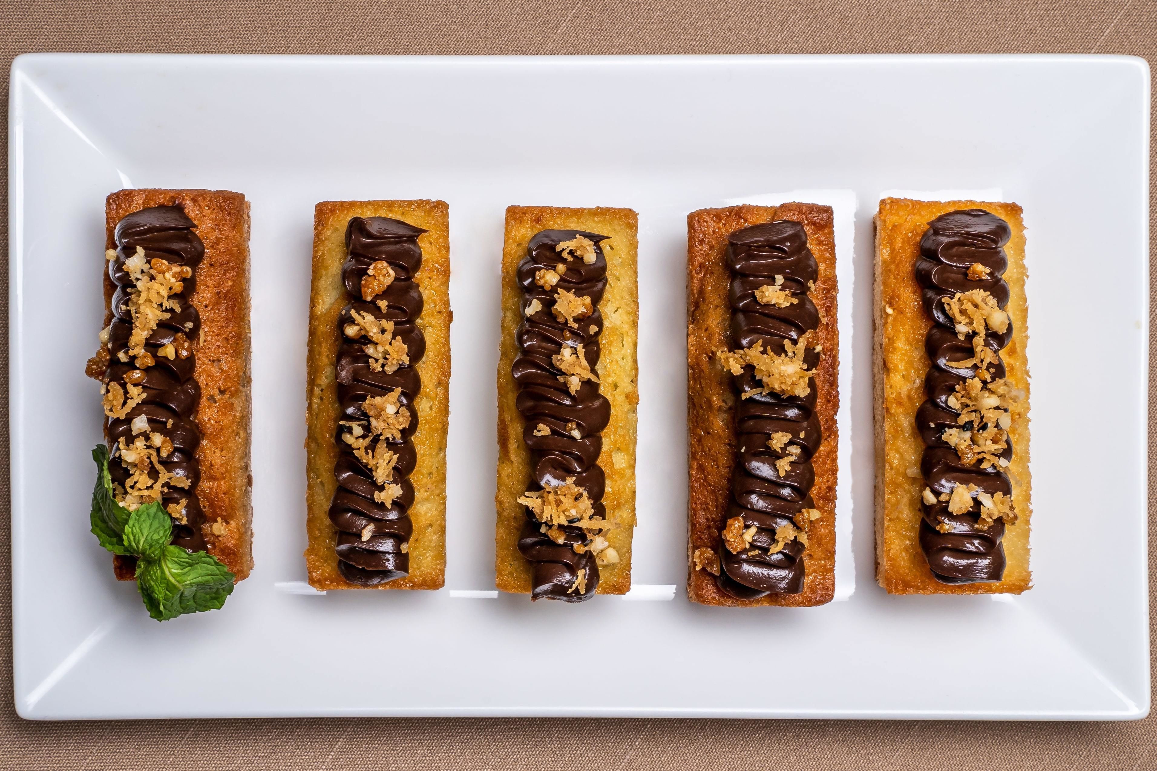 Cinq pâtisseries rectangulaires sur une assiette blanche, chacune garnie de chocolat et de noix concassées, avec une feuille de menthe sur le côté gauche.