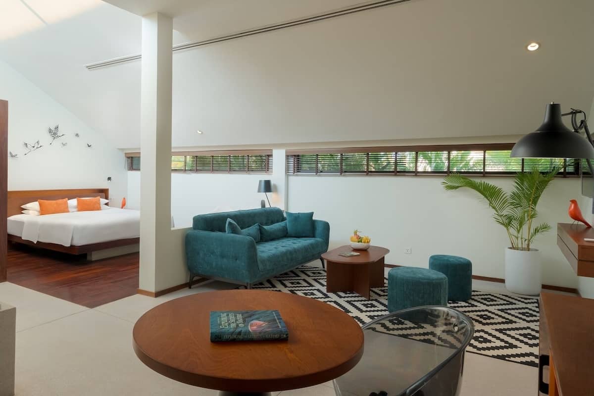 A modern hotel room with a bed, a sofa, a dining table, and a small kitchen area.