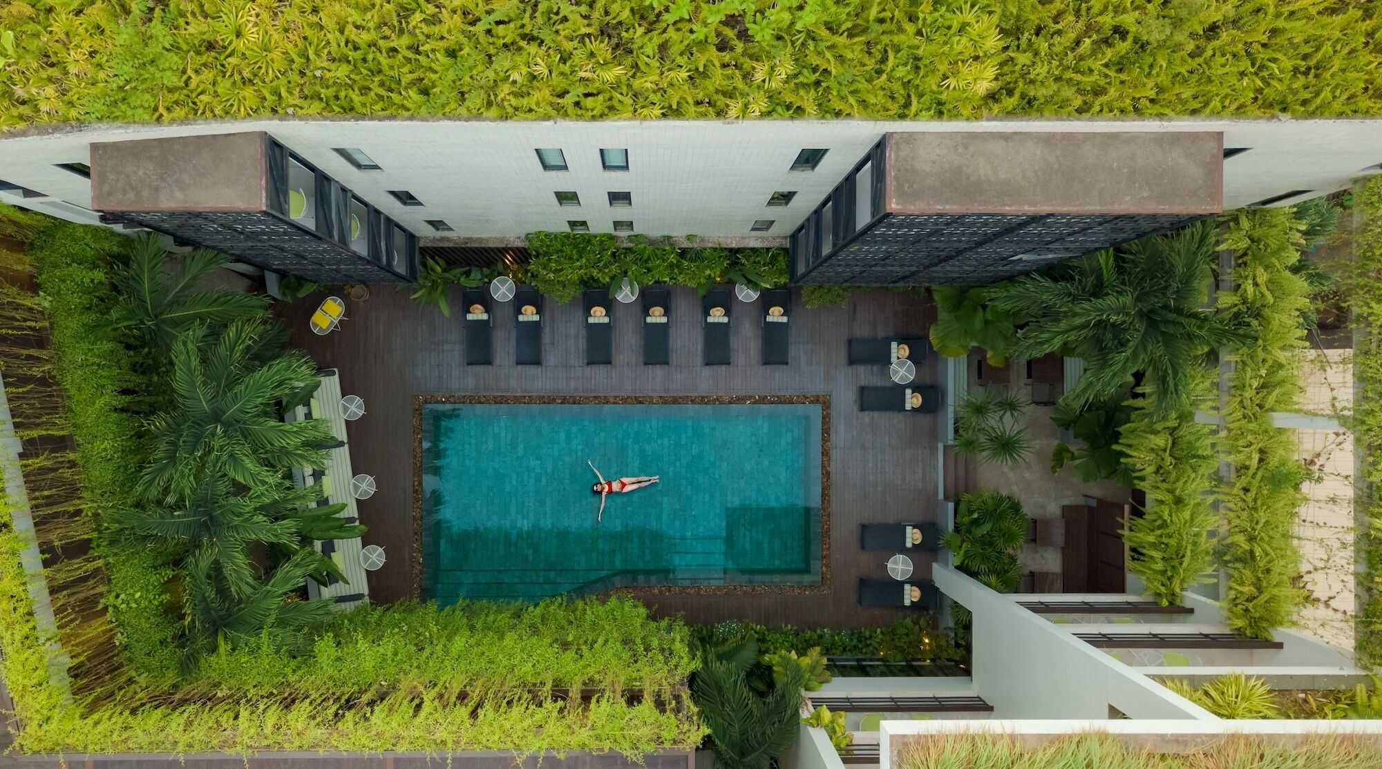 Et luftfoto viser et moderne hotel med en central swimmingpool, udendørs siddepladser og frodig beplantning.
