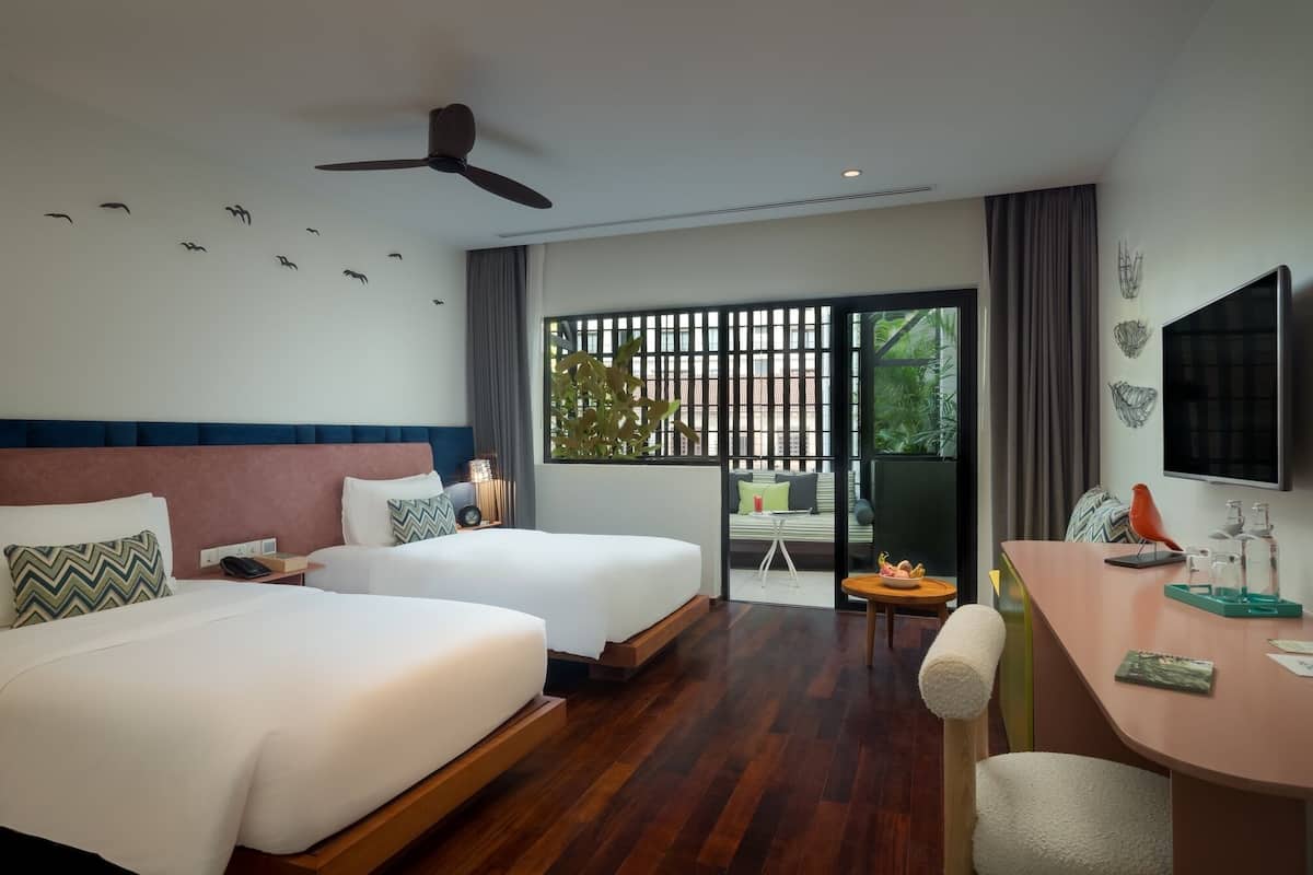 A hotel room with two beds, a desk, a chair, a TV, and a balcony with a view of greenery.