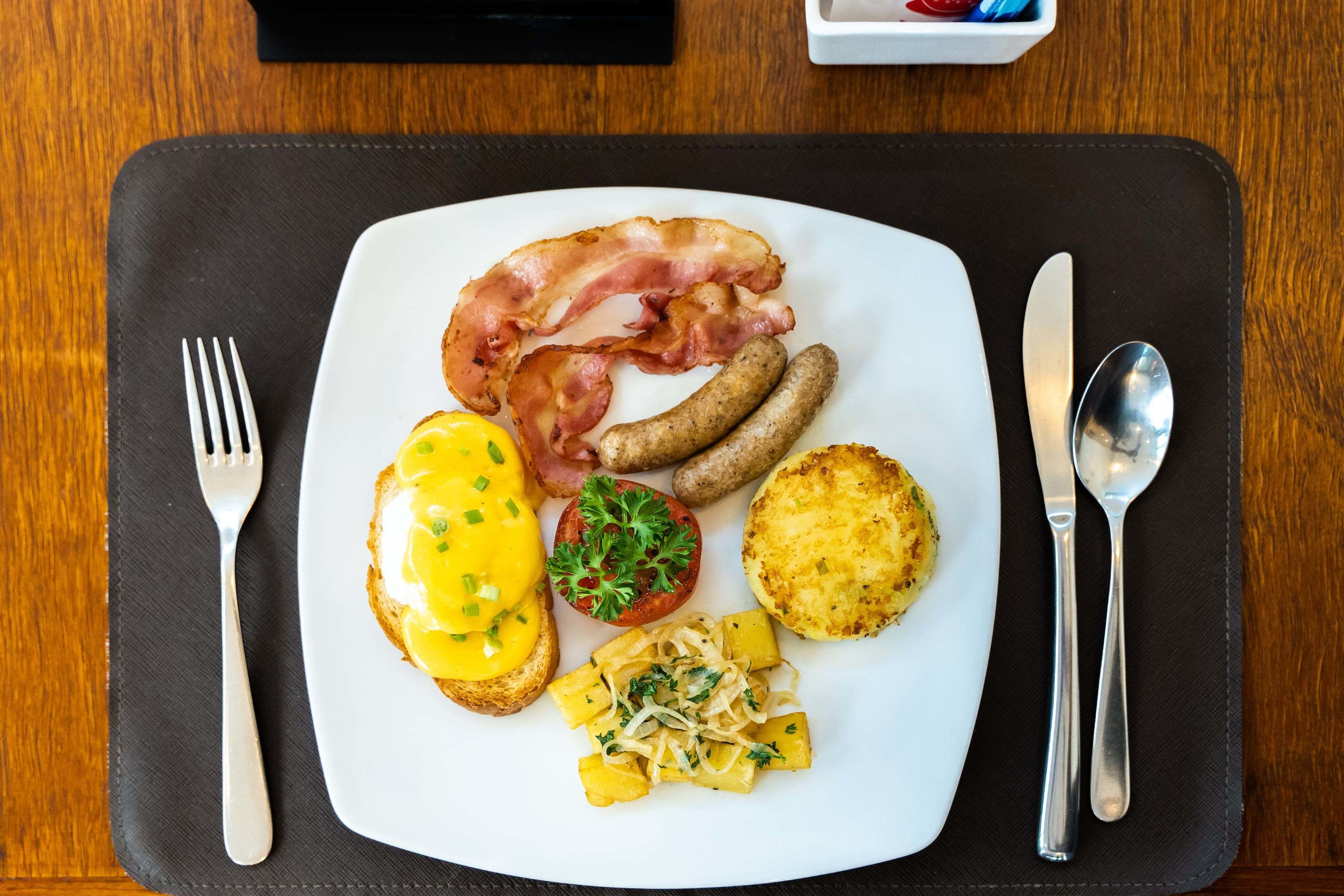A breakfast plate with bacon, sausages, eggs, tomatoes, and potatoes on a white plate.