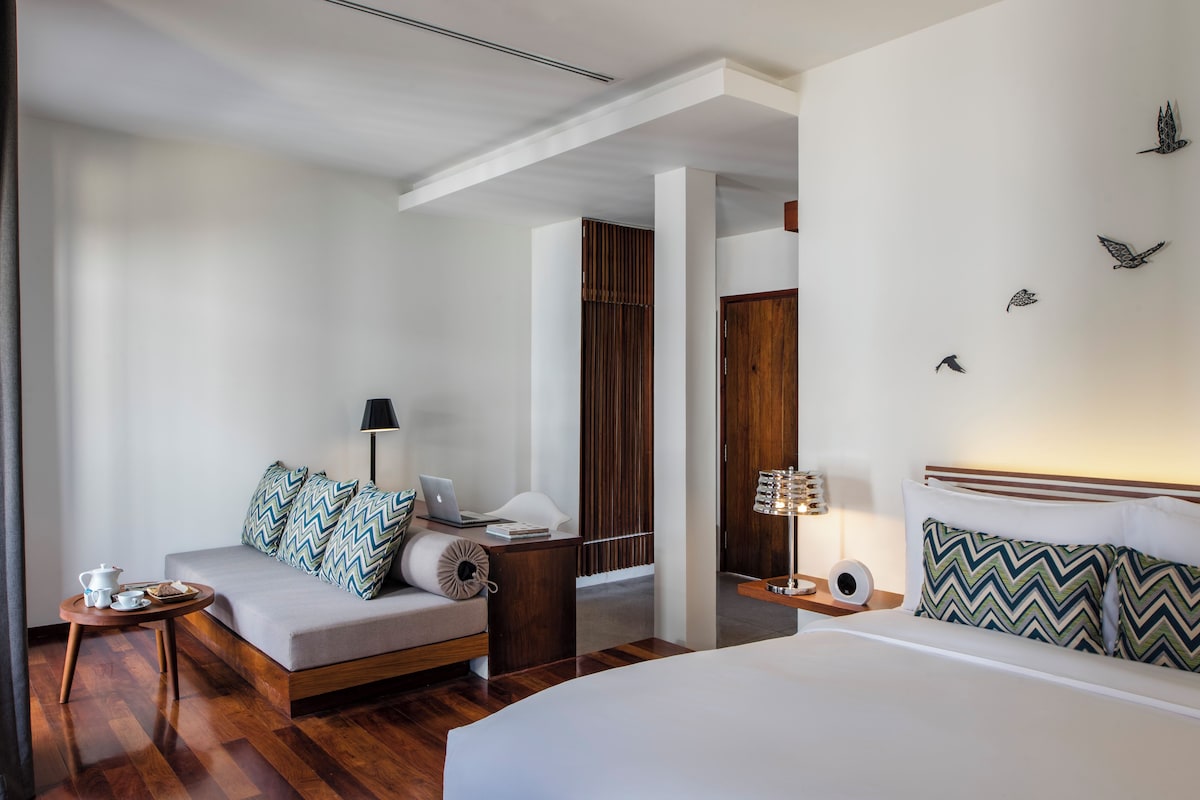 A modern hotel room with a bed, a sofa, a desk, and a nightstand.