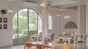 Uma ampla sala de jantar com mesas e cadeiras brancas, grandes janelas em arco e um lustre.