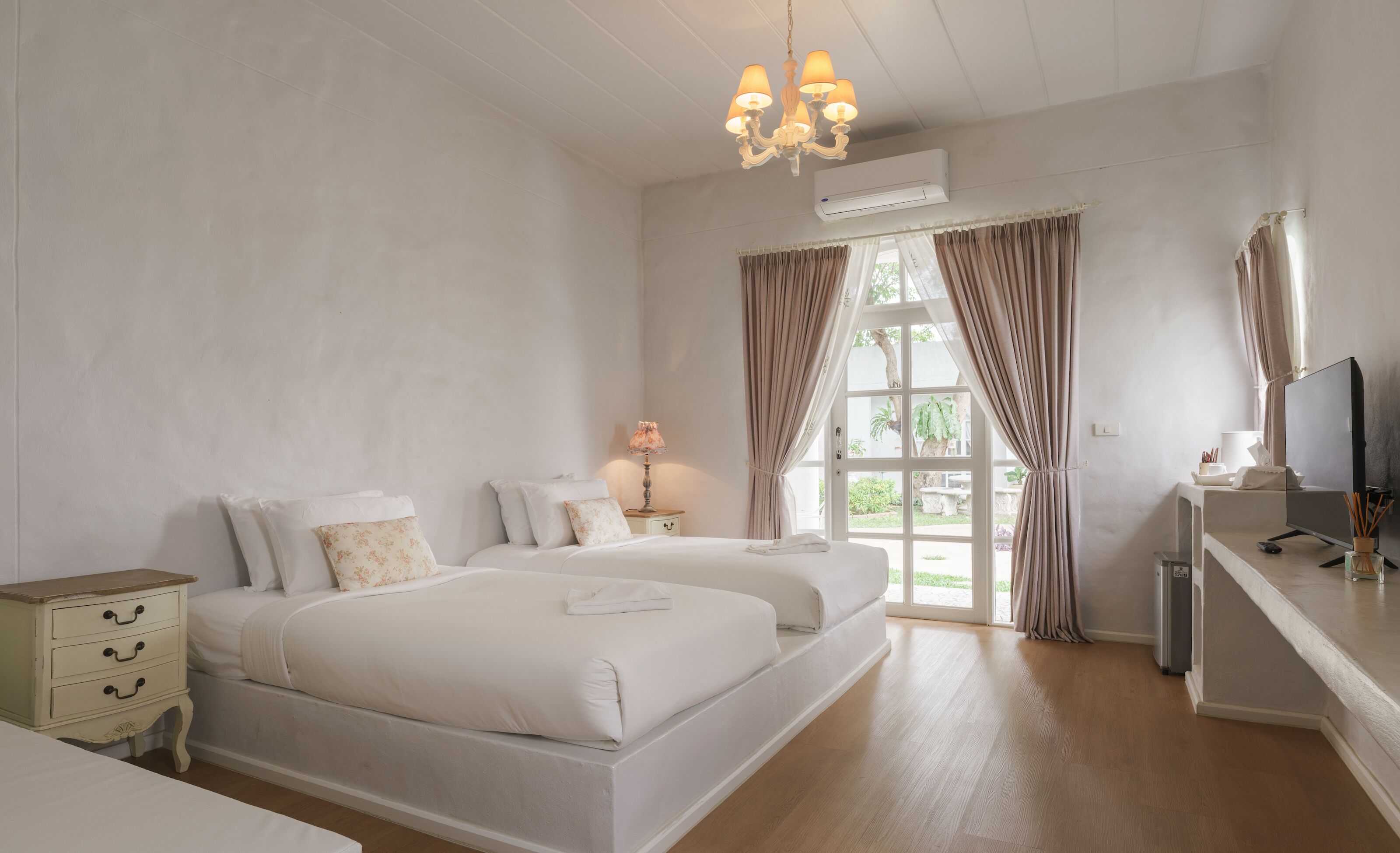 Ein Hotelzimmer mit zwei Betten, einem Schreibtisch mit Computer, einer Lampe und einem großen Fenster mit Vorhängen.