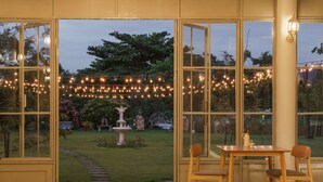 Quarto com uma grande janela com vista para um jardim, uma mesa de madeira com cadeiras e luzes de corda.