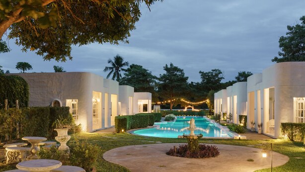 Um pátio com piscina, mesas de pedra e uma fonte, cercado por edifícios brancos e vegetação.