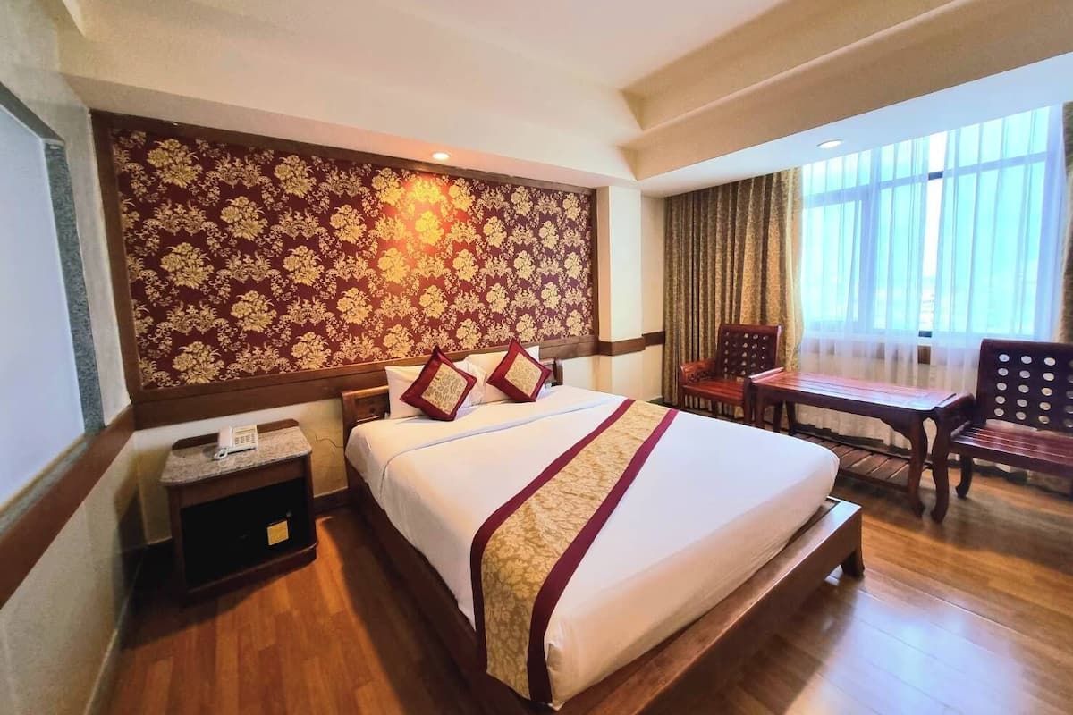 Quarto de hotel com uma cama grande, uma escrivaninha e uma cadeira. Há um telefone na escrivaninha e uma janela com cortinas.