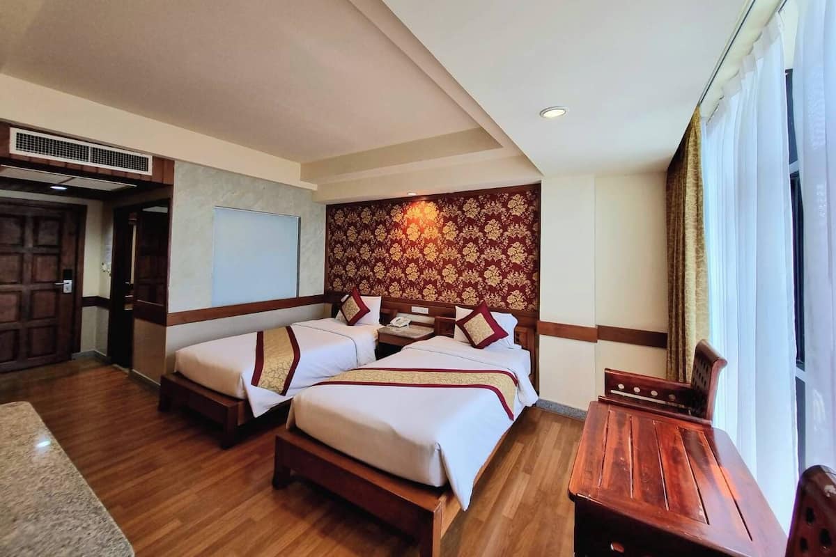 Quarto de hotel com duas camas, uma escrivaninha de madeira, uma janela grande com cortinas e um painel de cabeceira estampado.