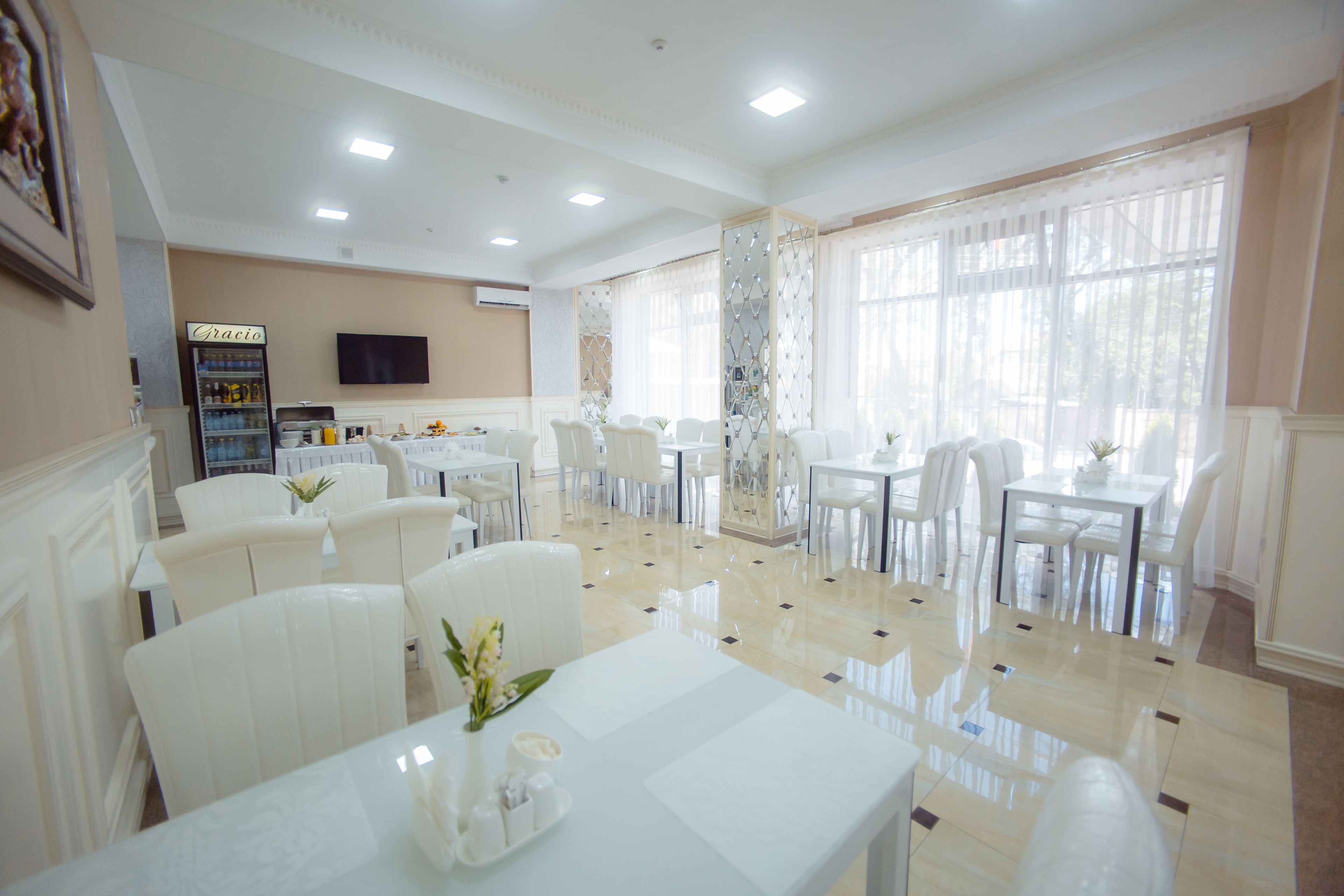 A hotel dining area with white furniture, a refrigerator, and a television on the wall.